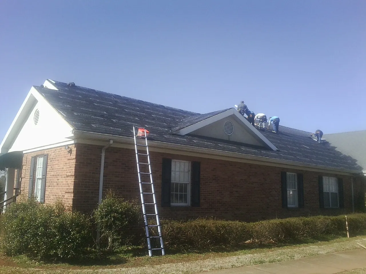 Crew tearing shingles off a brick ranch roof with a ladder set — Metal Roof Repair work by our crew in Harmon