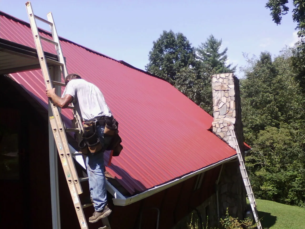Roofer on a ladder beside a red metal roof and stone chimney — Soffit & Fascia Repair by Roofing Brothers in Harmon